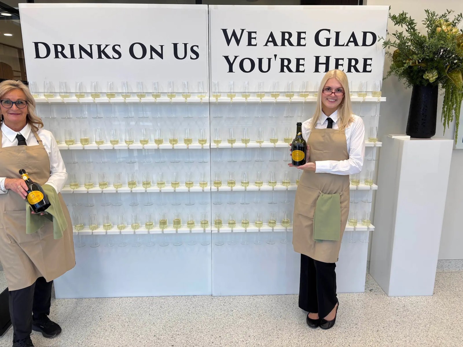 Two bartenders beside a champagne wall reading Drinks On Us