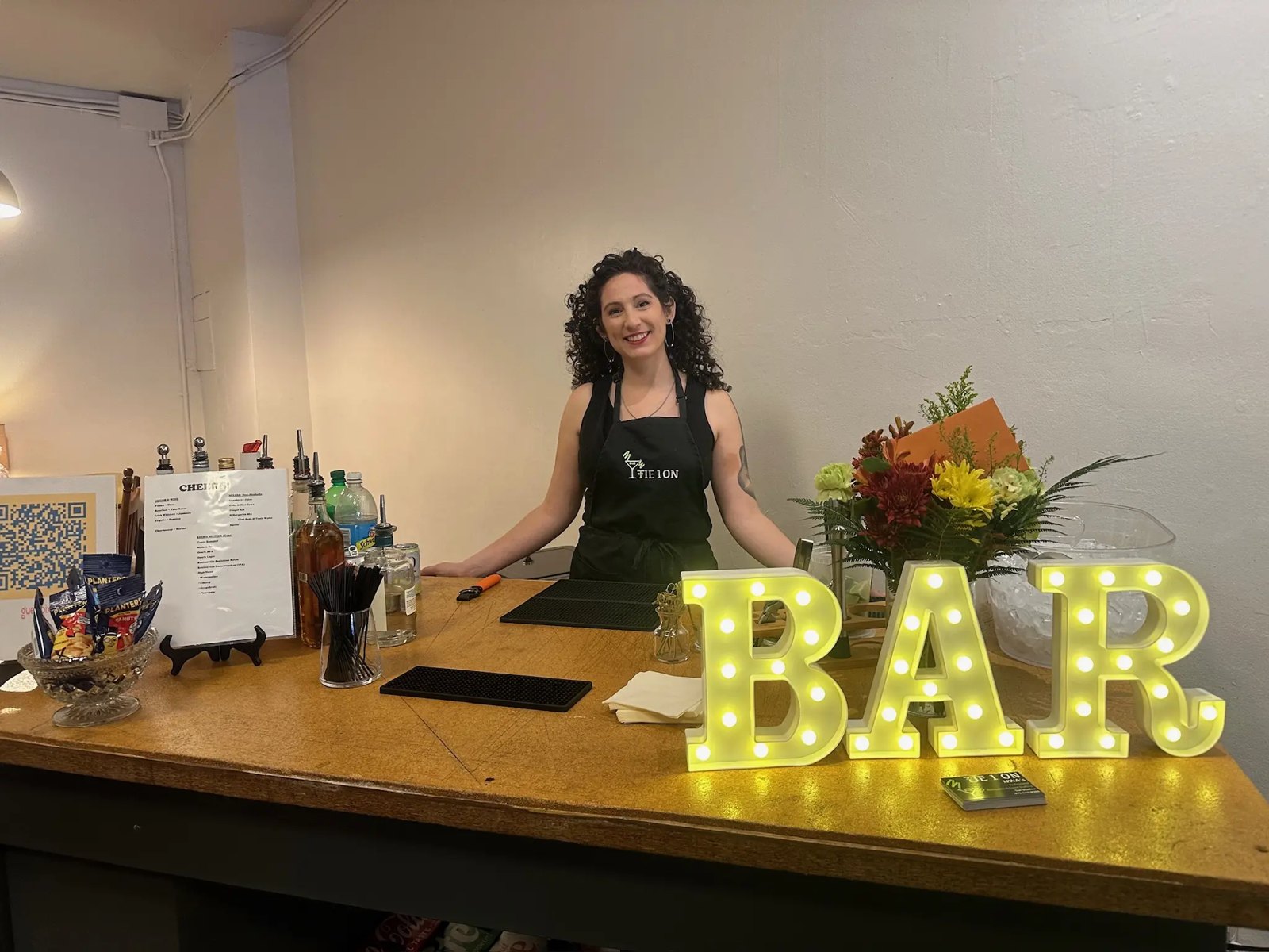 A bartender behind a home bar with a lit BAR marquee sign